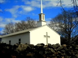 White church building and steeple against a blue sky behind a stone wall
